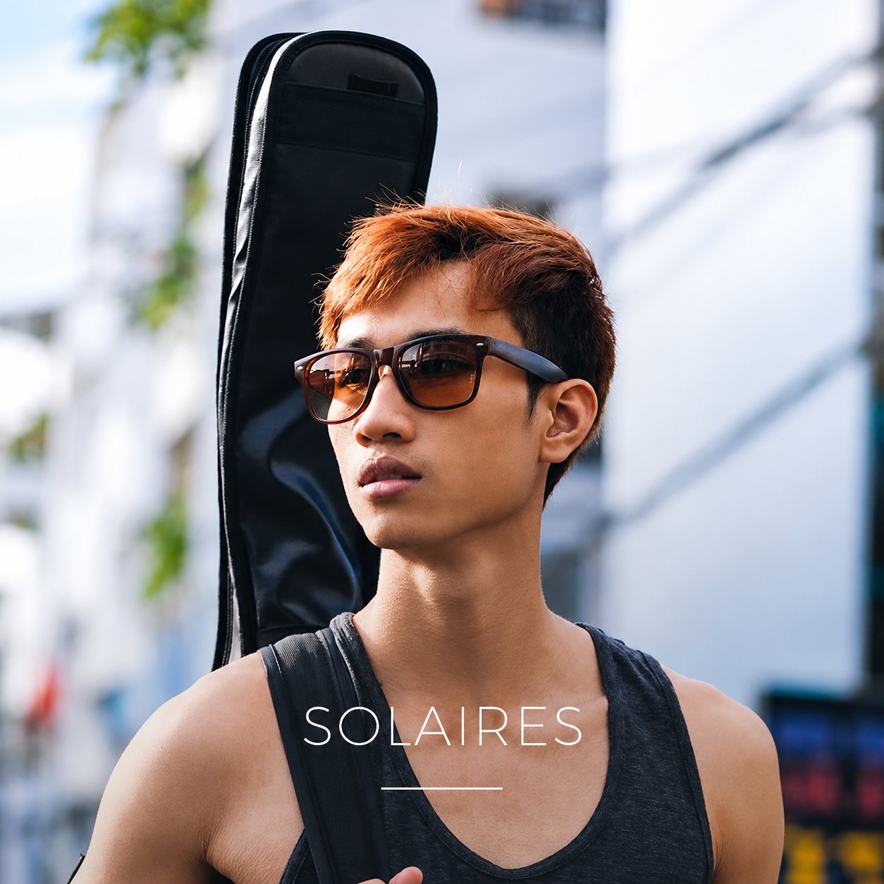 Un jeune homme en lunettes de soleil, une housse de guitare sur le dos, dans la rue.