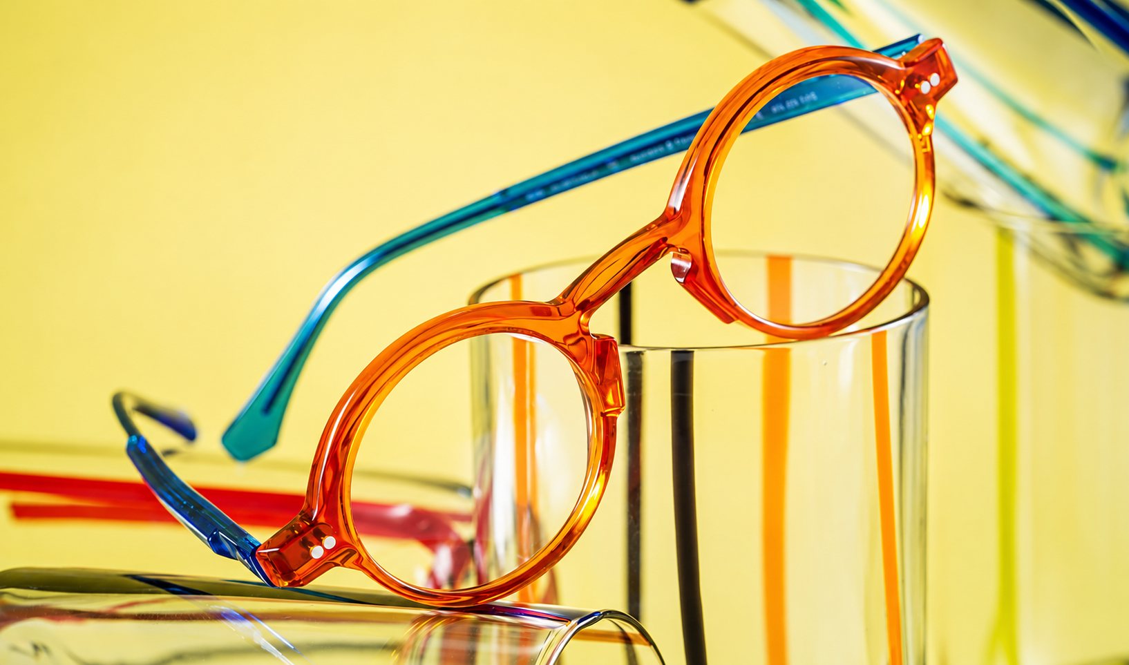 Une paire de lunettes rondes orange translucide aux branches bleues posée sur des verres rayés, devant un fond jaune.
